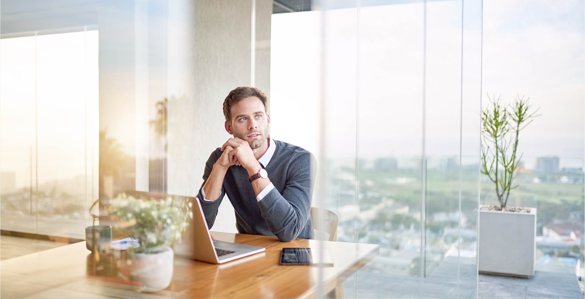 Mann sitzt nachdenklich am Schreibtisch mit Laptop in einem modernen Büro mit Glasfassade, symbolisch für nachhaltige Finanzberatung.