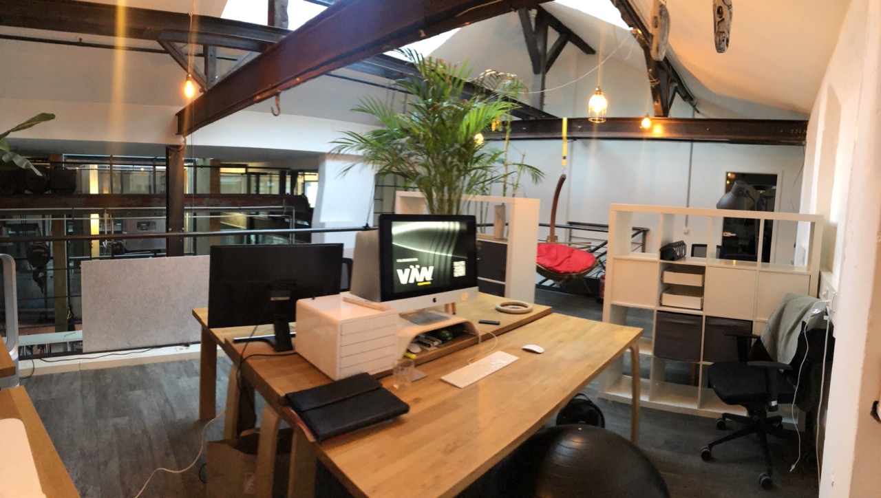 Modernes Designbüro mit Holzschreibtischen, Pflanzen, iMac-Arbeitsplatz und industriellen Dachbalken im Loftstil.